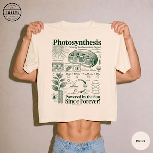 Pode incluir: Uma t-shirt de cor marfim com uma impressão gráfica verde-escura que detalha o processo da fotossíntese. O design inclui diagramas científicos de um cloroplasto, uma planta, equações químicas e o texto Photosynthesis Turning Sunbeams into Sugar! Powered by the Sun Since Forever!