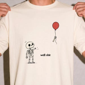 May include: A cream-coloured t-shirt featuring a black line-art illustration of a skeleton reaching towards a floating red balloon held by a detached skeletal hand. The text "well shit" is printed in black lowercase letters next to the skeleton.