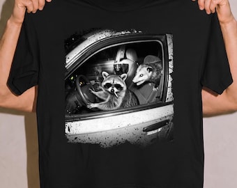 May include: A black t-shirt featuring a graphic print of a raccoon wearing sunglasses and holding a cigarette while sitting in the driver's seat of a car, with an opossum in the passenger seat and a skunk visible in the back seat of the vehicle.