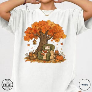 May include: A white t-shirt featuring a graphic of a cat sitting in a green armchair under an autumn tree, reading a book. The scene includes a birdhouse, a basket of pumpkins, a steaming mug, and fallen orange leaves. The logo "NUMBER TWELVE STUDIOS" is printed in the bottom left corner.