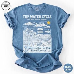 May include: A blue jean coloured t-shirt featuring a white graphic print of the water cycle. The design includes text reading THE WATER CYCLE, Same Water Since the Dinosaurs, and Powered by the Rain Since Forever, along with scientific diagrams.