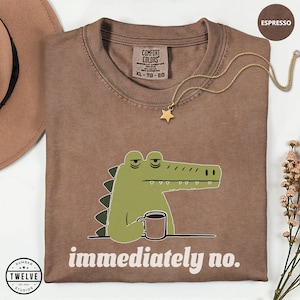 May include: An espresso-colored t-shirt featuring a graphic of a green crocodile holding a coffee mug with a tired expression. Below the crocodile, the text reads "immediately no." The shirt has a Comfort Colors label at the inner neckline.
