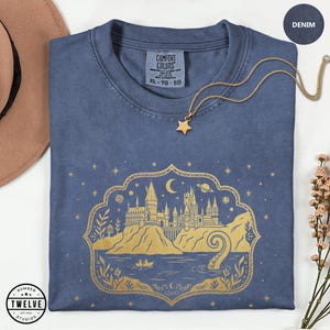 May include: Denim blue t-shirt with a gold graphic of a castle, moon, and stars. A gold star pendant necklace rests on the shirt. The shirt has a tag that says "Comfort Colors" and the size is XL-TG-EG. The shirt is from Twelve Studios.