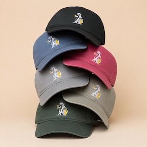 May include: A stack of six baseball caps in black, blue, red, gray, olive, and dark green. Each cap features an embroidered graphic of a white dinosaur holding a yellow cocktail glass and a yellow suitcase, centered on the front panel of the crown.