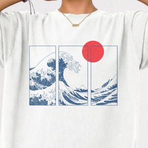 May include: A white t-shirt featuring a graphic print divided into three vertical rectangular panels. The design displays a blue line art illustration of a large ocean wave, a distant mountain, and a solid red circle representing the sun.