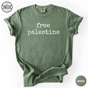 Pode incluir: Uma t-shirt de manga curta verde-musgo com decote redondo. A peça apresenta o texto branco "free palestine" impresso numa fonte estilo máquina de escrever no peito. O tecido tem uma textura ligeiramente desbotada e estilo vintage.