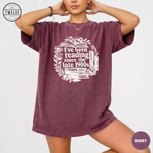 Puede incluir: Una camiseta de manga corta color baya con un estampado gráfico blanco de libros apilados y flores. El texto dice, I've been reading since the late 1900s. La camiseta tiene un corte relajado con cuello redondo y un acabado lavado vintage.