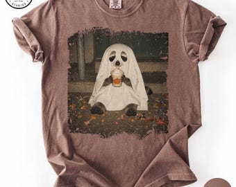 May include: A brown crew neck t-shirt featuring a graphic of a raccoon dressed as a ghost, holding a coffee cup with a pumpkin design. The graphic is set against a dark, textured background with scattered sweets. The shirt has a vintage, washed-out look.