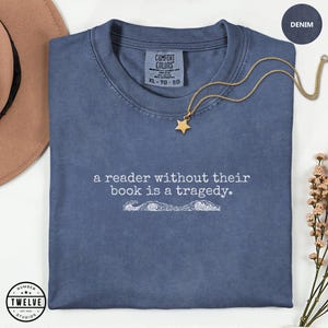 Op de afbeelding: Denimblauwe Comfort Colors T-shirt met de tekst "a reader without their book is a tragedy." Een gouden sterhanger hangt aan een delicate ketting. Het shirt is netjes opgevouwen, met een bruine hoed en bloemen op de achtergrond.