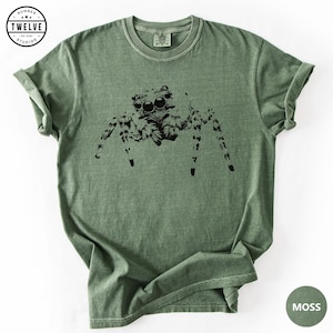 Puede incluir: Una camiseta de manga corta color verde musgo con un estampado gráfico negro de una araña saltarina en el pecho. La prenda tiene cuello redondo y una textura visible en la tela. Un logotipo circular en la esquina superior izquierda dice NUMBER TWELVE STUDIOS.