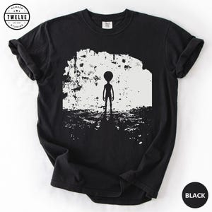 Op de afbeelding: Een zwart t-shirt met korte mouwen en een witte grafische print van een staand alien-silhouet tegen een textuurrijke, cirkelvormige achtergrond. Het cirkelvormige logo linksboven bevat de tekst Number Twelve Studios.