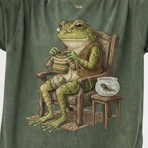 May include: A sage green t-shirt featuring a graphic illustration of a green frog wearing round glasses and a plaid vest, sitting in a wooden chair while knitting a small sweater with yarn, with a tadpole in a glass bowl on a side table.
