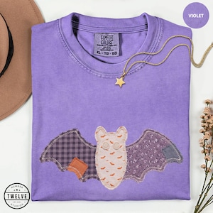 Op de afbeelding: Een violet t-shirt met een centrale vleermuisapplicatie gemaakt van diverse stoffen, waaronder een crèmekleurig lijfje met kleine oranje vleermuisjes, een paarse ruitjesvleugel en een bloemrijke vleugel. Een gouden sterrenketting ligt op de kraag.
