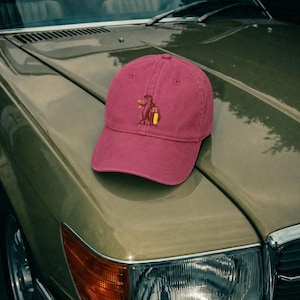 Può includere: Un cappellino da baseball rosa scuro con un ricamo che raffigura una lontra marrone che tiene in mano un bicchiere da cocktail giallo e una valigia gialla. Il cappellino presenta una corona strutturata, una visiera curva e un bottone.
