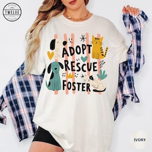 Könnte beinhalten: Ein elfenbeinfarbenes Kurzarm-T-Shirt mit einem Grafikdruck und dem Text ADOPT RESCUE FOSTER. Das Design enthält Illustrationen von zwei Hunden und zwei Katzen in Gelb-, Blaugrün- und Schwarztönen vor vertikalen rosa Streifen.