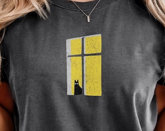 May include: A charcoal grey t-shirt featuring a graphic print of a yellow window frame with a distressed texture. A small, solid black cat silhouette with two white circular eyes is positioned at the bottom of the window frame, looking outward.