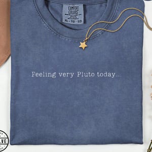 Może przedstawiać: Niebieski t-shirt z okrągłym dekoltem i białym napisem Feeling very Pluto today... na piersi. Koszulka posiada metkę marki Comfort Colors przy wewnętrznym dekolcie i jest zestawiona ze złotym naszyjnikiem z zawieszką w kształcie gwiazdki.