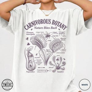 Puede incluir: Camiseta blanca con una ilustración botánica detallada de plantas carnívoras, incluyendo una Venus atrapamoscas y un rocío del sol. El diseño incluye etiquetas científicas y el texto "CARNIVOROUS BOTANY Nature Bites Back". La camiseta tiene cuello redondo y mangas cortas.