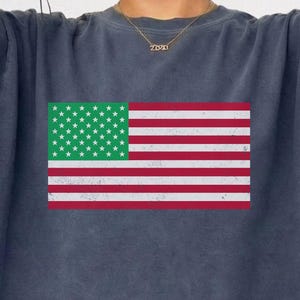 Könnte beinhalten: Ein dunkelblaues Sweatshirt mit Rundhalsausschnitt und einem verwaschenen Aufdruck der amerikanischen Flagge mit grünem Feld und weißen Sternen, kombiniert mit einer goldfarbenen Kette mit dem Schriftzug 2023.