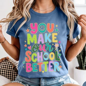 Könnte beinhalten: Ein blaues Kurzarm-T-Shirt mit dem mehrfarbigen Schriftzug "YOU MAKE OUR SCHOOL BETTER" in einer verspielten Schriftart. Das Design enthält kleine grafische Elemente wie Smileys, Sterne, Herzen und eine Blume, die um den Text verteilt sind.