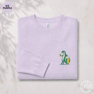 Puede incluir: Una sudadera de cuello redondo color violeta hielo con un gráfico bordado en el pecho izquierdo de un lagarto verde que sostiene una copa de cóctel amarilla y se apoya en una maleta amarilla con ruedas. La prenda tiene cuello y puños acanalados.