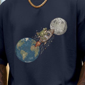 Könnte beinhalten: Ein marineblaues T-Shirt mit einer grafischen Illustration eines grünen Frosches, der auf einer Rakete von der Erde in Richtung Mond fliegt. Die Rakete hat orangefarbene Flammen und das Design enthält kleine Sterne im Hintergrund.
