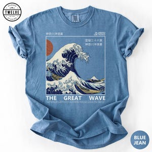 May include: A blue jean colored t-shirt featuring a graphic print of The Great Wave off Kanagawa. The design includes a red sun, Japanese text, coordinates, and the text THE GREAT WAVE. A logo for Number Twelve Studios is in the top left corner.