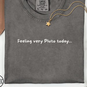 Może przedstawiać: Ciemnoszary t-shirt z białym napisem "Feeling very Pluto today..." nadrukowanym z przodu. Złoty naszyjnik z małą zawieszką w kształcie gwiazdki spoczywa na dekolcie koszulki, która posiada metkę marki Comfort Colors.