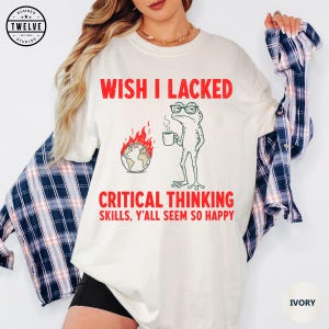 May include: An ivory t-shirt featuring a graphic of a green frog wearing glasses and holding a coffee mug next to a burning globe. The red text reads: WISH I LACKED CRITICAL THINKING SKILLS, Y'ALL SEEM SO HAPPY. The shirt is layered over a navy plaid shirt.