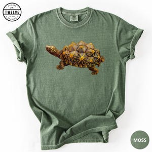 Puede incluir: Una camiseta de manga corta color verde musgo con un gráfico detallado de una tortuga con un caparazón geométrico y texturizado en tonos marrón y dorado. La esquina superior izquierda muestra un logotipo circular con el texto Number Twelve Studios Est. 2008.