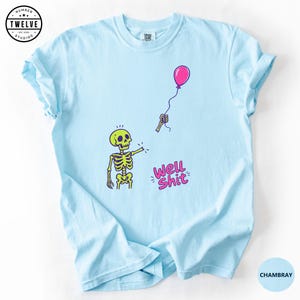 May include: A light blue t-shirt featuring a graphic of a yellow skeleton with a detached arm holding a pink balloon. The text "WELL SHIT" is printed in pink below the skeleton. The design includes small motion lines around the skeleton's head.