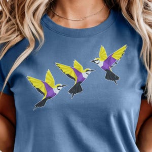 Könnte beinhalten: Ein blaues T-Shirt mit Rundhalsausschnitt und einem Grafikdruck von drei fliegenden Vögeln. Jeder Vogel ist in einem geometrischen Low-Poly-Stil mit leuchtenden gelben, violetten und weißen Farbblöcken dargestellt, die diagonal angeordnet sind.