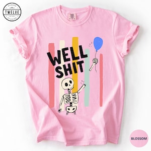 May include: A light pink t-shirt featuring a graphic of a skeleton holding a blue balloon. The text WELL SHIT is printed in black above the skeleton. Behind the graphic are vertical stripes in shades of coral, cream, yellow, and teal.