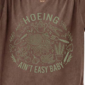 Peut inclure: Un t-shirt marron avec un motif circulaire imprimé en vert clair. Le design représente des outils de jardinage, un panier de légumes et des fleurs, accompagné du texte "HOEING AIN'T EASY BABY" disposé en arc autour de l'illustration.