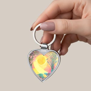 May include: Heart-shaped keychain with a vibrant, colorful landscape design. The artwork features a yellow sun, a winding path, and a small bird, set against a backdrop of pink and green foliage. The keychain has a silver metal finish.