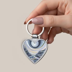 May include: Heart-shaped keychain with a silver metal frame. The artwork inside depicts a landscape with a large, bright sphere in the center, surrounded by swirling colors of blue, gray, and yellow. The keychain is held by a hand.