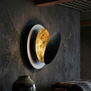 May include: Contemporary wall sconce with a circular design. The sconce has a white base, a textured gold-coloured centre, and a black, curved accent. It's mounted on a dark grey textured wall, creating a contrast of light and shadow.