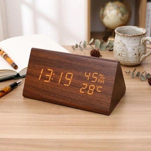 May include: A wooden triangular digital clock showing the time as 13:19, 45% humidity, and 28 degrees Celsius. The clock has a dark brown wood grain finish. The clock is on a light brown surface.