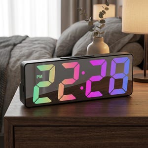 May include: Black digital alarm clock displaying "22:28 PM" with a gradient of colors. The clock sits on a wooden nightstand. A lamp and vase with decorative stems are in the background. The clock has a sleek, modern design.