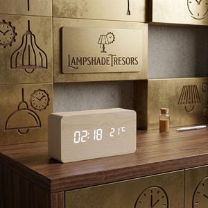 May include: A light-colored wooden digital clock displaying the time 02:18 and the temperature 21°C. The clock sits on a dark wood surface with a brass-colored background featuring the text "LAMPSHADE TRESORS".