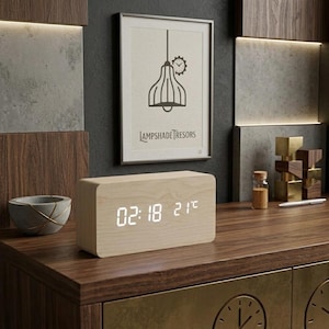 Peut inclure: Une horloge numérique en bois clair affichant l'heure et la température. L'horloge est posée sur une surface en bois foncé, avec une impression encadrée d'un design de lampe. L'impression indique "LAMPSHADE TREASORS".