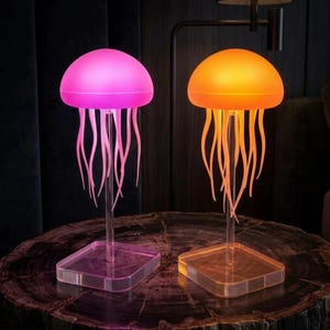 Könnte beinhalten: Zwei quallenförmige Lampen mit klarem Sockel und Stiel. Eine Lampe leuchtet rosa, die andere orange. Die Lampen haben eine abgerundete Oberseite und lange, fließende Tentakel. Die Lampen stehen auf einer dunklen, strukturierten Oberfläche.