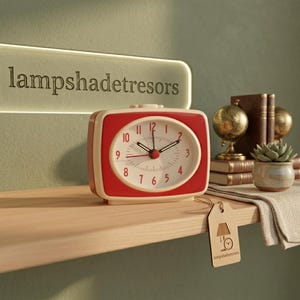 Puede incluir: Un reloj despertador retro rojo y crema con una esfera blanca. El reloj tiene números y manecillas negras y está colocado en una estantería de madera. El texto "lampshadetresors" es visible encima del reloj.