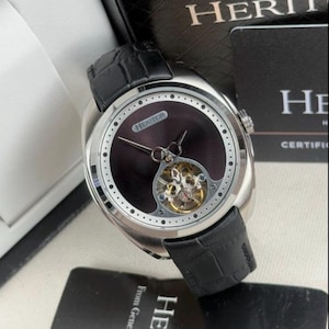 Könnte beinhalten: Eine silberne HERITOR-Uhr mit schwarzem Lederarmband. Das Zifferblatt der Uhr ist dunkel mit sichtbarem mechanischem Uhrwerk und Markennamen. Die Uhr wird in einer schwarzen Box mit sichtbarem Markennamen präsentiert.