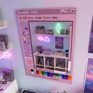 Könnte beinhalten: Ein pinkfarbener Wandkunstrahmen, der als Retro-Computerfenster gestaltet ist, mit dem Text "untitled - Paint". Im Inneren zeigen Regale Vintage-Spielkonsolen, Bücher und Neonkunst. Unten befindet sich eine Farbpalette.