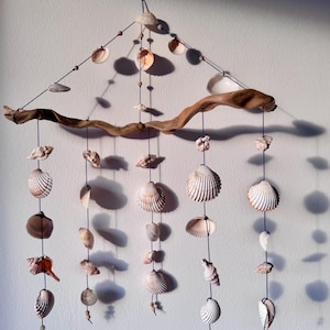Può includere: Mobile di conchiglie fatto a mano. Presenta una base in legno di deriva e fili neri. Varie conchiglie bianche, beige e marroni sono infilate, creando un'estetica costiera. Piccole perline di legno aggiungono dettagli.