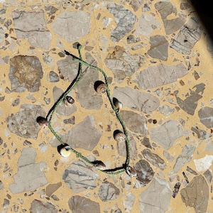 May include: A handmade necklace featuring small seashells strung on a green cord. The shells are a mix of white, tan, and brown, and the cord is knotted to secure the shells. The necklace is laid on a speckled, light-colored surface.