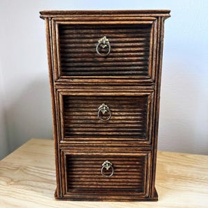 May include: A dark brown wooden chest with three horizontal drawers, each featuring a ribbed texture on the front panel and a small, circular brass-toned metal pull handle centered on the drawer face with a decorative floral-shaped backplate.