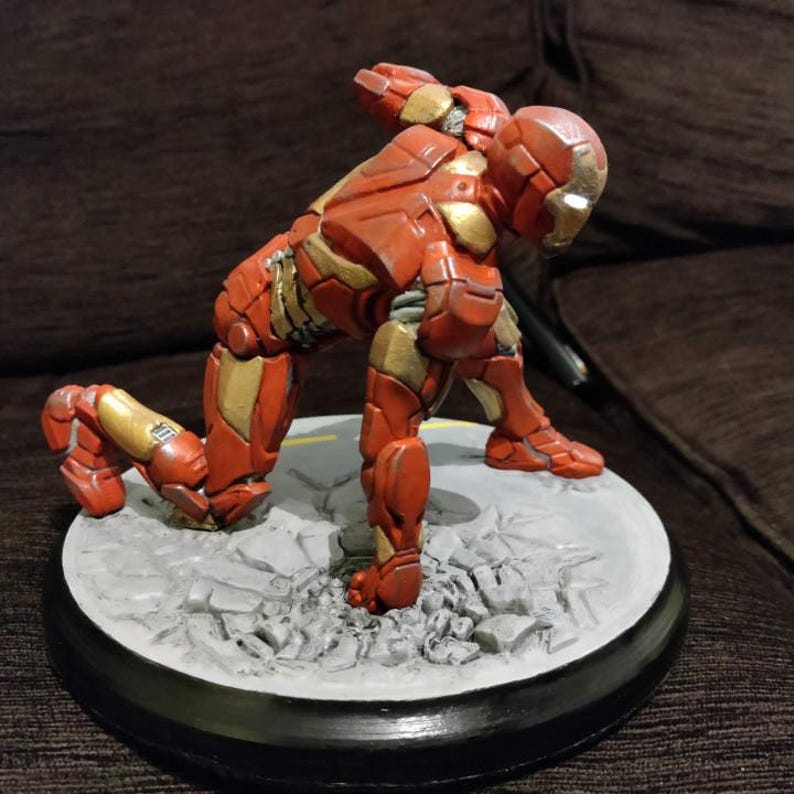 May include: A red and gold Iron Man figurine in a crouching pose. The figurine is on a gray circular base with a black border, and the base has a cracked concrete design. The figure is highly detailed.