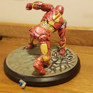 May include: A red and gold Iron Man figurine in a dynamic pose, set on a round, textured base. The figurine's armor features detailed paneling and a metallic sheen. A small, detached piece lies near the base.